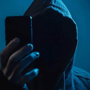 A person in a hooded sweatshirt holds up a smartphone, with their face obscured by darkness and a digital pixelated effect, creating an anonymous, mysterious appearance. Blue lighting enhances the dramatic mood, hinting at Stolen Device Protection.