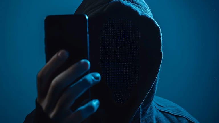 A person in a hooded sweatshirt holds up a smartphone, with their face obscured by darkness and a digital pixelated effect, creating an anonymous, mysterious appearance. Blue lighting enhances the dramatic mood, hinting at Stolen Device Protection.