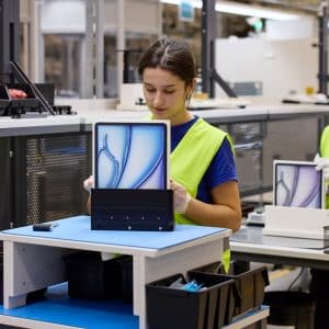 Two workers in a factory, part of an Apple global sourcing strategy, assemble tablets wearing yellow safety vests. One places a tablet with a blue abstract screen into packaging, while another works at a similar station in the background.