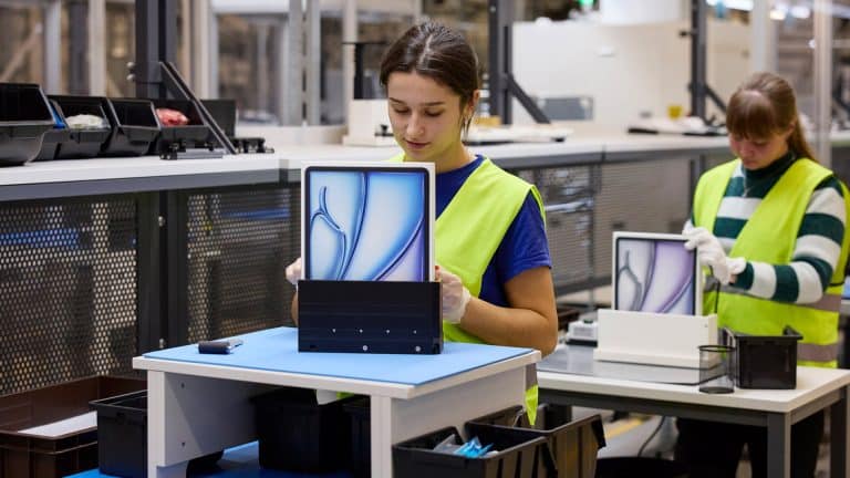 Two workers in a factory, part of an Apple global sourcing strategy, assemble tablets wearing yellow safety vests. One places a tablet with a blue abstract screen into packaging, while another works at a similar station in the background.