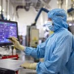 A person wearing a blue cleanroom suit, mask, and gloves operates a touchscreen monitor in a high-tech laboratory, reflecting the precision and efficiency characteristic of the Apple global sourcing strategy.