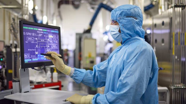 A person wearing a blue cleanroom suit, mask, and gloves operates a touchscreen monitor in a high-tech laboratory, reflecting the precision and efficiency characteristic of the Apple global sourcing strategy.