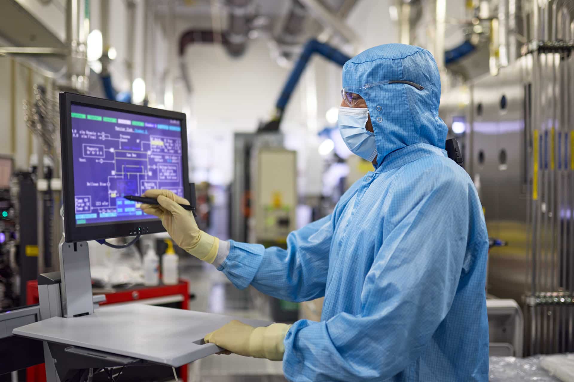 A person wearing a blue cleanroom suit, mask, and gloves operates a touchscreen monitor in a high-tech laboratory, reflecting the precision and efficiency characteristic of the Apple global sourcing strategy.