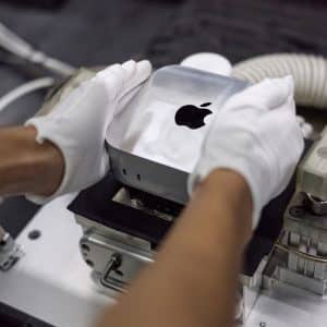 A person wearing white gloves places a silver Apple Mac mini on a machine, possibly for assembly or testing, reflecting Apple's global sourcing strategy in a manufacturing or laboratory setting.