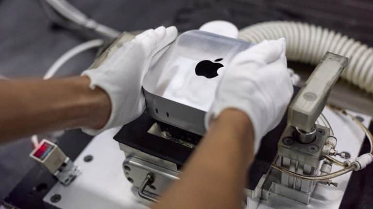 A person wearing white gloves places a silver Apple Mac mini on a machine, possibly for assembly or testing, reflecting Apple's global sourcing strategy in a manufacturing or laboratory setting.