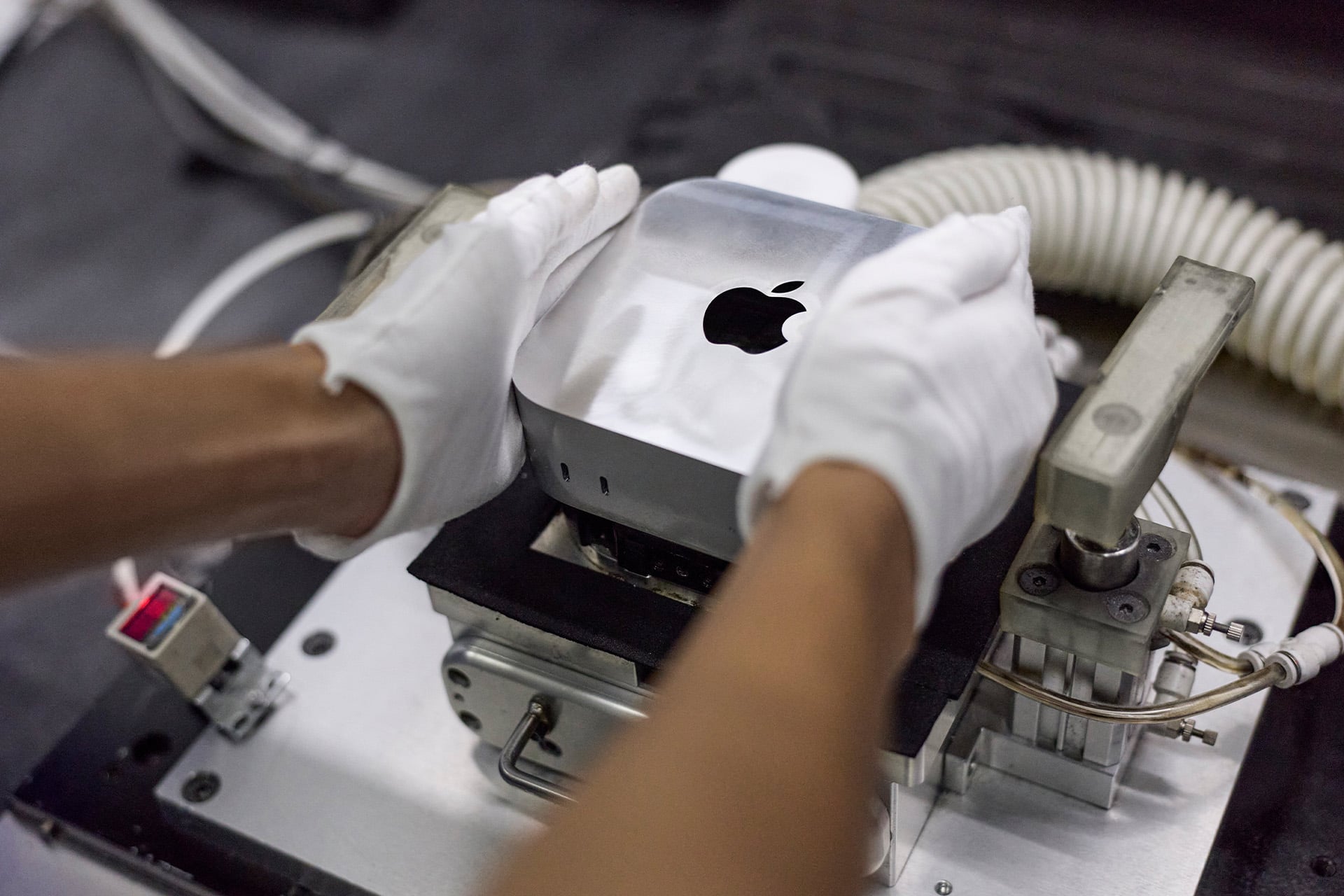 A person wearing white gloves places a silver Apple Mac mini on a machine, possibly for assembly or testing, reflecting Apple's global sourcing strategy in a manufacturing or laboratory setting.