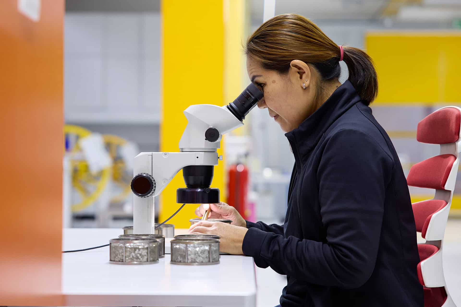 A woman sits at a laboratory table looking through a microscope, applying precision akin to Apple global sourcing strategy. She examines specimens in small dishes while bright yellow and orange panels light the background.
