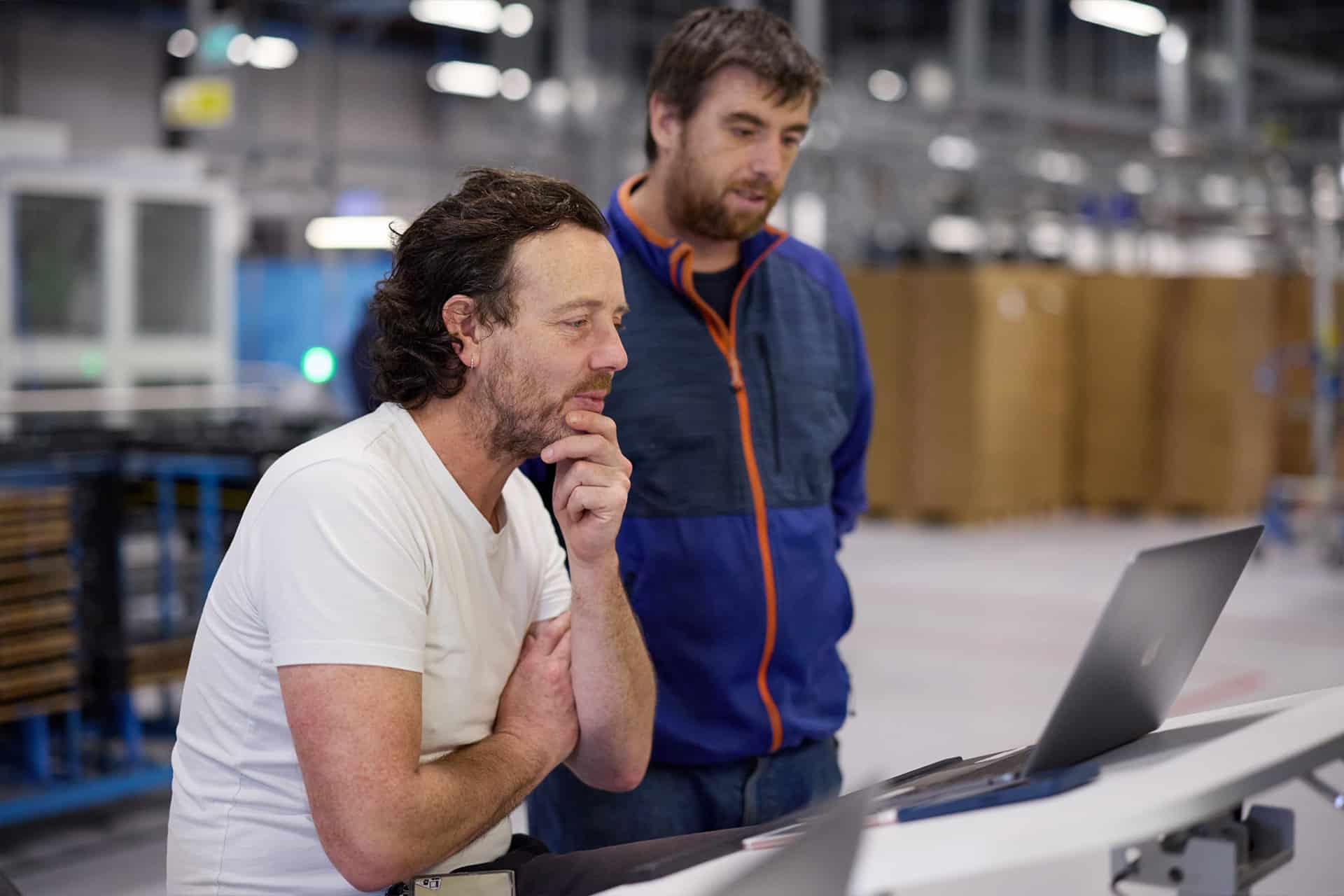 Two men stand at a workstation in an industrial setting, focused on a laptop screen as they discuss the Apple global sourcing strategy. One wears a white T-shirt, the other a blue jacket, with shelves and equipment in the background.