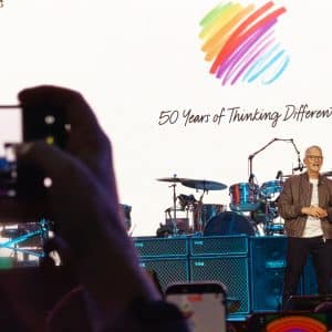 A person stands on stage in front of drums and equipment, with a colorful heart logo and "50 Years of Thinking Different" on a large screen behind them. Audience members snap photos, capturing the moment before returning to off-screen time.
