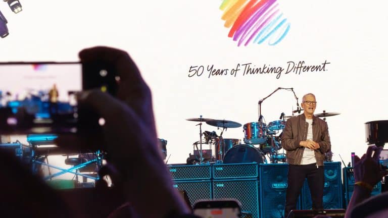 A person stands on stage in front of drums and equipment, with a colorful heart logo and "50 Years of Thinking Different" on a large screen behind them. Audience members snap photos, capturing the moment before returning to off-screen time.