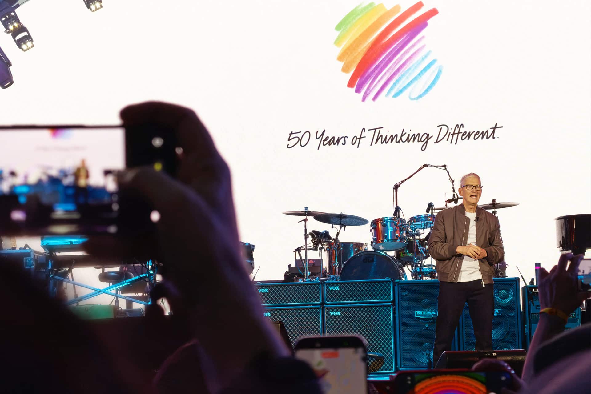 A person stands on stage in front of drums and equipment, with a colorful heart logo and "50 Years of Thinking Different" on a large screen behind them. Audience members snap photos, capturing the moment before returning to off-screen time.