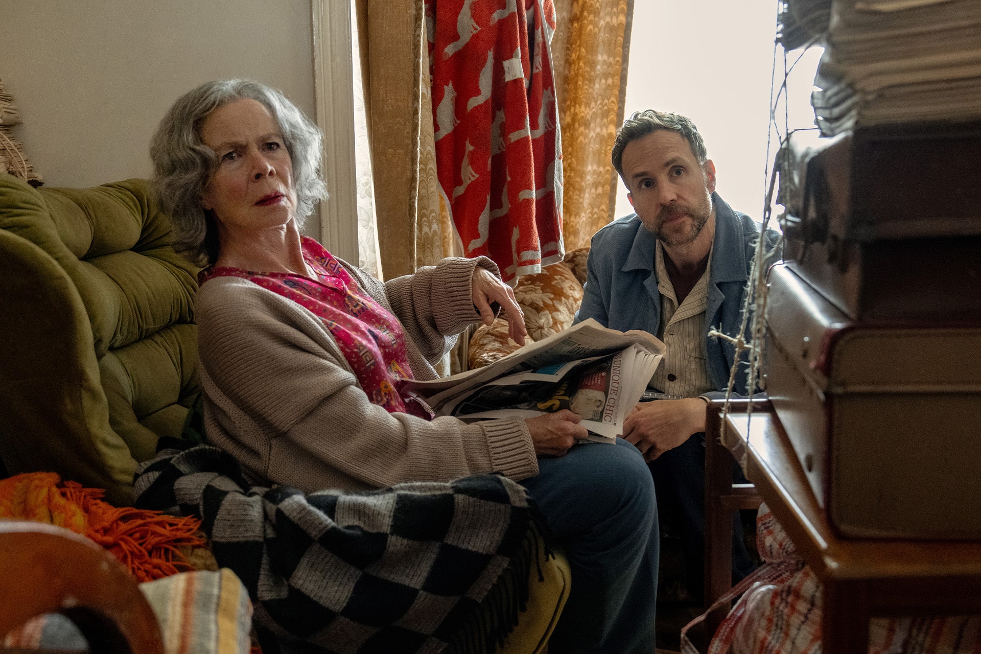 An older woman with gray hair and a man with short hair sit in a cluttered, warmly lit room. The woman looks surprised while holding a newspaper featuring Trying Season 5, as the man beside her gazes intently forward.