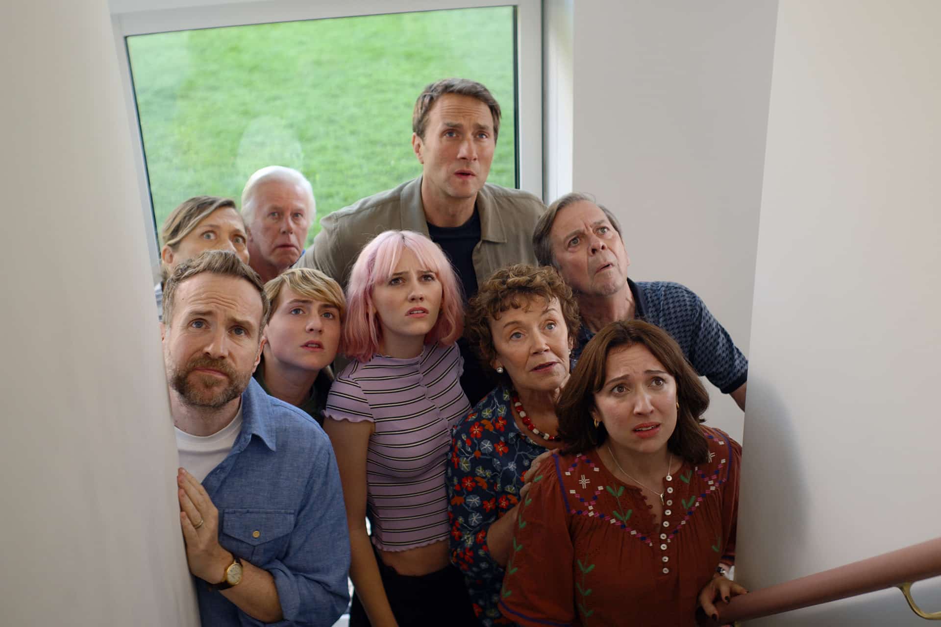 A group of eight people, including men and women of various ages, stand closely together on a staircase, looking up with anxious and concerned expressions—a scene reminiscent of the tension in Trying Season 5. A large window behind them shows grass outside.