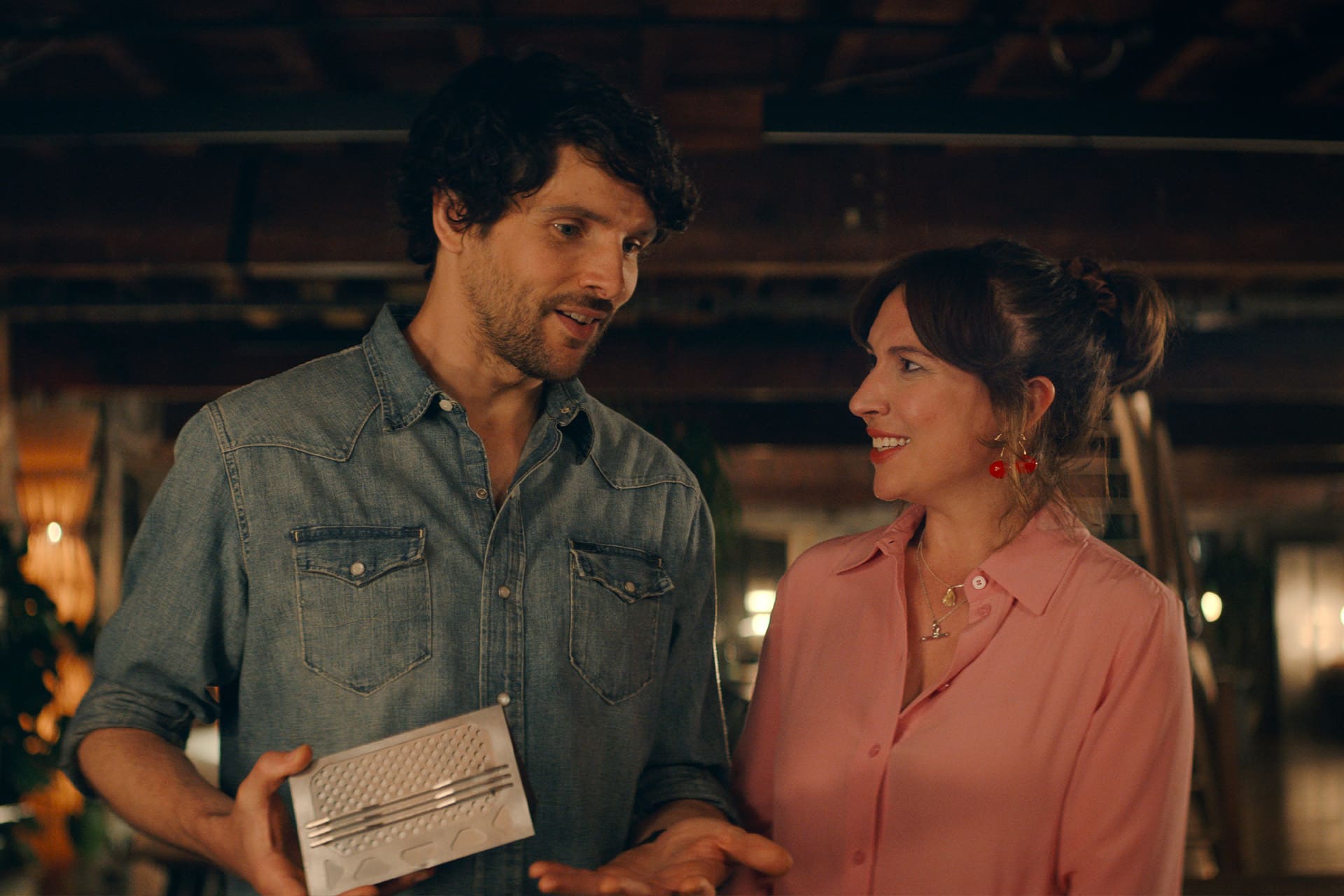 A man in a denim shirt holds a small white gift box while standing next to a woman in a pink blouse with red earrings. Indoors, they smile at each other warmly, capturing the heartfelt moments fans love in Trying Season 5.