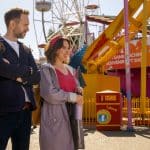 A man and woman stand smiling at an amusement park with colorful rides, a Ferris wheel, and a souvenir shop in the background—ready for adventure just like in Trying Season 5. The woman wears a headband and raincoat; the man carries a backpack.
