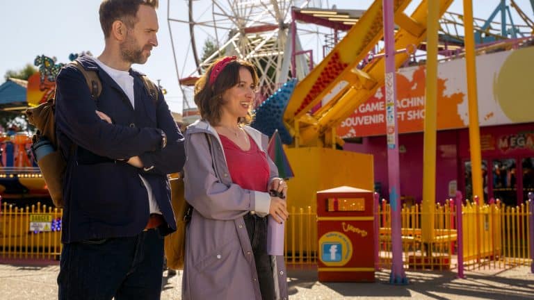 A man and woman stand smiling at an amusement park with colorful rides, a Ferris wheel, and a souvenir shop in the background—ready for adventure just like in Trying Season 5. The woman wears a headband and raincoat; the man carries a backpack.