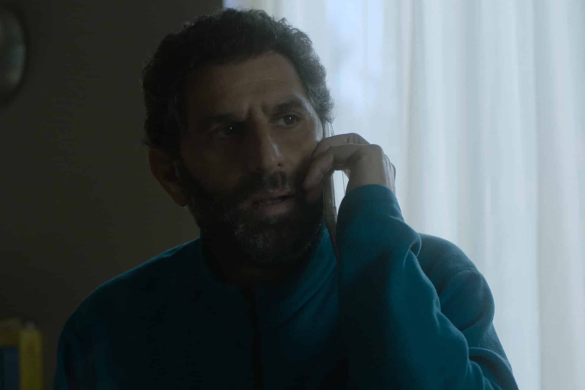 A man with curly dark hair and a beard, wearing a teal fleece jacket, is talking on a phone. He looks concerned and is sitting in a dimly lit room with light coming through sheer curtains behind him, resembling a scene from the Unconditional series.