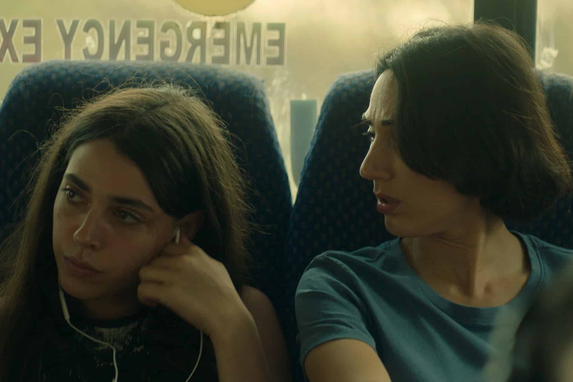 Two women sit closely on a bus in this scene from the Unconditional series; one gazes out the window with earphones in, appearing upset, while the other looks at her with concern. "EMERGENCY EXIT" is visible on the window behind them.