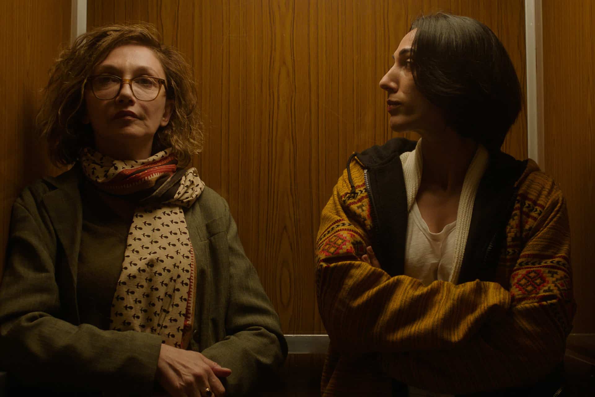 Two women stand in a wood-paneled elevator in the Unconditional series. One wears glasses, a scarf, and a jacket, gazing forward; the other, with short dark hair and a patterned jacket, looks sideways at her companion. Both appear thoughtful.