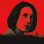 A woman’s face, tinted red and black, dominates the image against a bold red background. Part of the Unconditional series, two small silhouetted figures walk along a fence below, creating a dramatic contrast.
