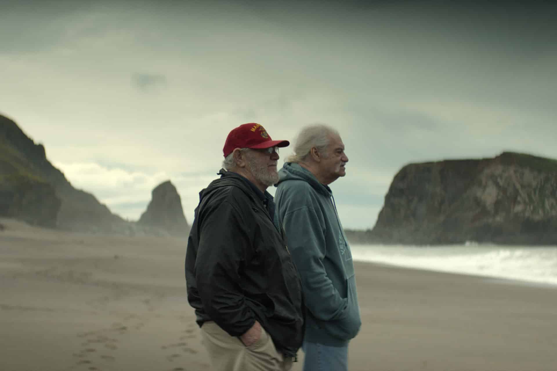 Two older men stand side by side on a deserted beach, gazing thoughtfully toward the ocean. With rocky cliffs and cloudy skies behind them, their scene recalls the reflective tone seen in recent Apple TV nominations for the 86th Annual Peabody Awards.