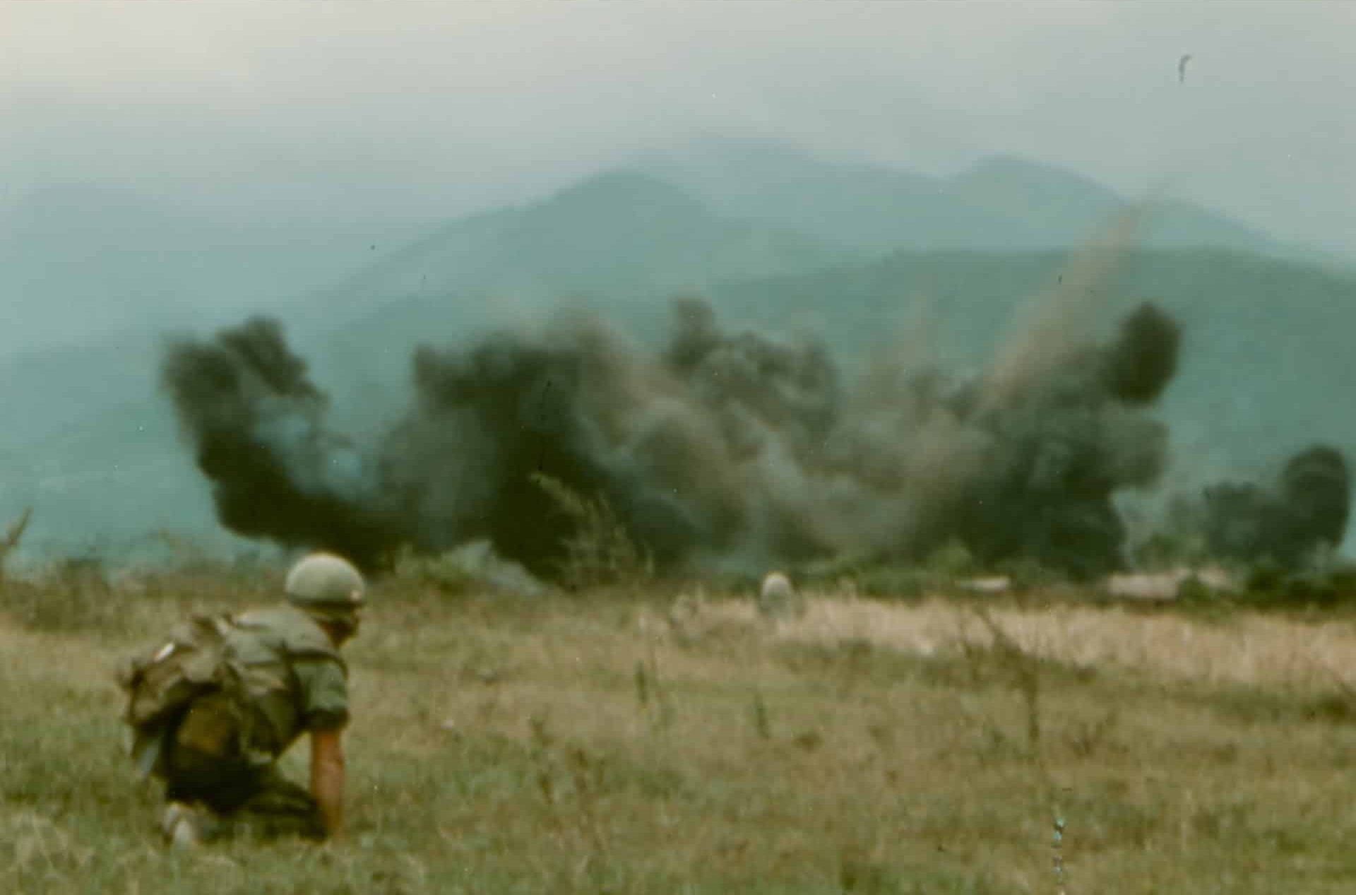A soldier crouches in a grassy field as a large explosion erupts, sending smoke and debris skyward—a scene reminiscent of BAFTA wins for outstanding war cinematography, with mountains looming under a cloudy sky.