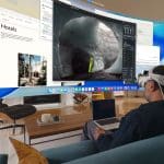A man wearing a VR headset sits on a couch with a laptop, using Vision Pro Mac Display to view multiple large virtual screens showcasing travel and nature content in a modern living room.