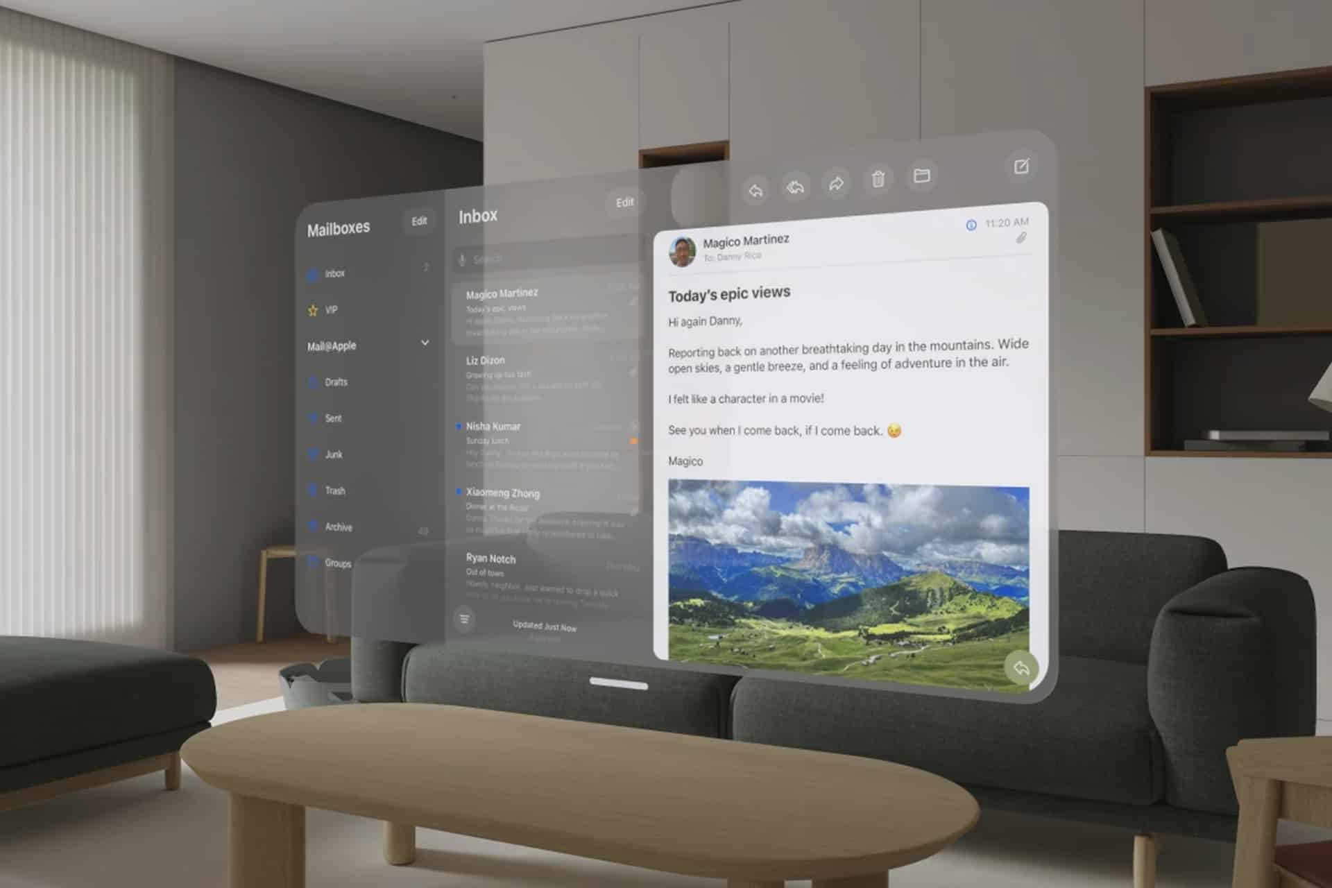 A person uses Vision Pro Mac Display to view a virtual email inbox projected in their living room, featuring a message about mountain views with a landscape photo. The room has a sofa, table, and minimalist decor.