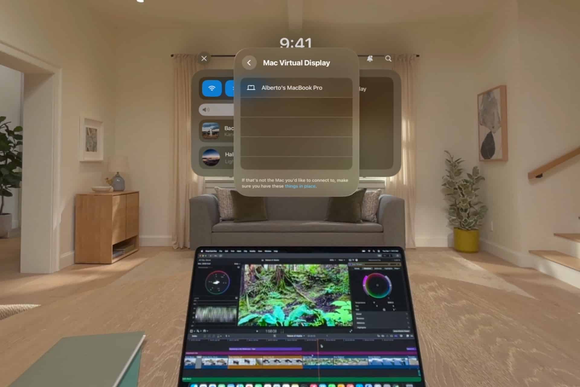 A person using a Vision Pro headset sees a digital display floating above a laptop running video editing software, with a modern living room in the background, seamlessly blending Mac Display capabilities with virtual reality.