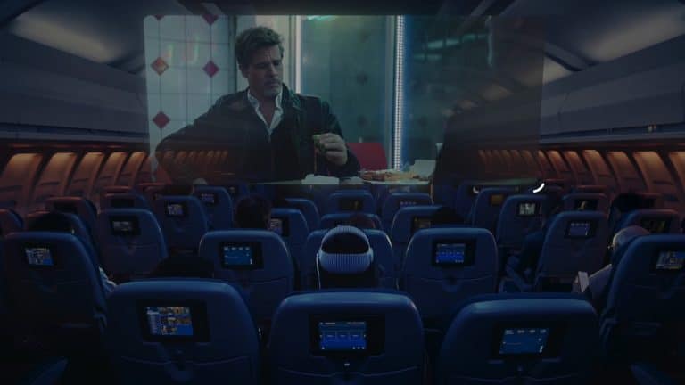 Passengers watch a movie on a large screen at the front of a dimly lit airplane cabin, with individual seatback screens and the option to use Apple Vision Pro travel mode for an immersive in-flight entertainment experience.