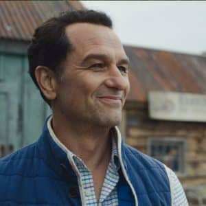 A man with short dark hair, wearing a blue vest over a plaid shirt, smiles while standing outside in front of weathered wooden buildings with metal roofs—a scene reminiscent of Apple TV Widow’s Bay.