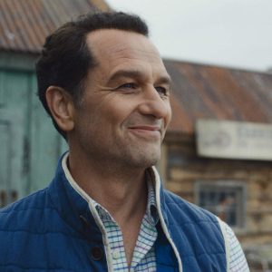 A man with short dark hair, wearing a blue vest over a plaid shirt, smiles while standing outside in front of weathered wooden buildings with metal roofs—a scene reminiscent of Apple TV Widow’s Bay.