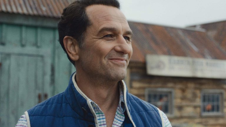 A man with short dark hair, wearing a blue vest over a plaid shirt, smiles while standing outside in front of weathered wooden buildings with metal roofs—a scene reminiscent of Apple TV Widow’s Bay.