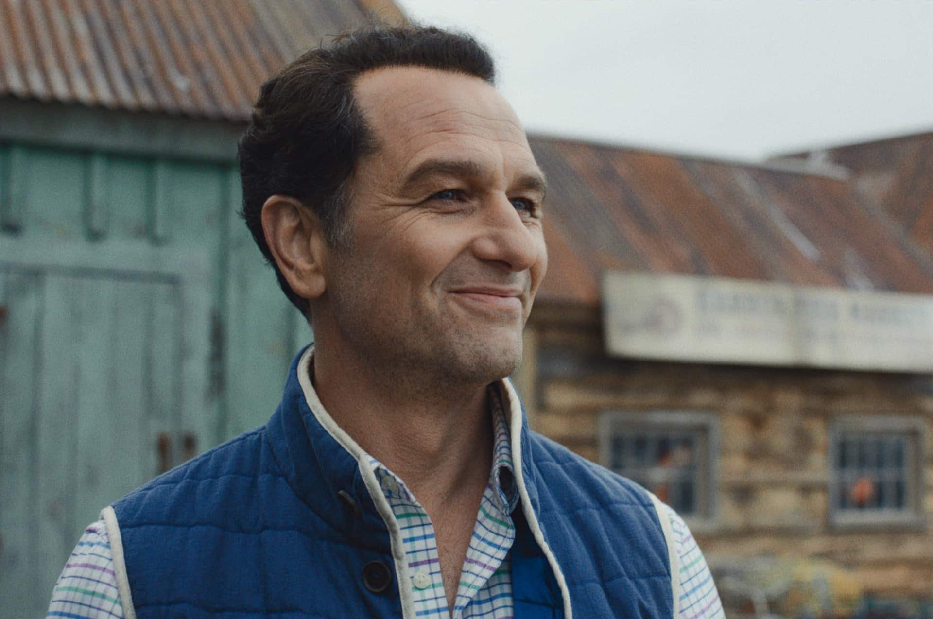 A man with short dark hair, wearing a blue vest over a plaid shirt, smiles while standing outside in front of weathered wooden buildings with metal roofs—a scene reminiscent of Apple TV Widow’s Bay.