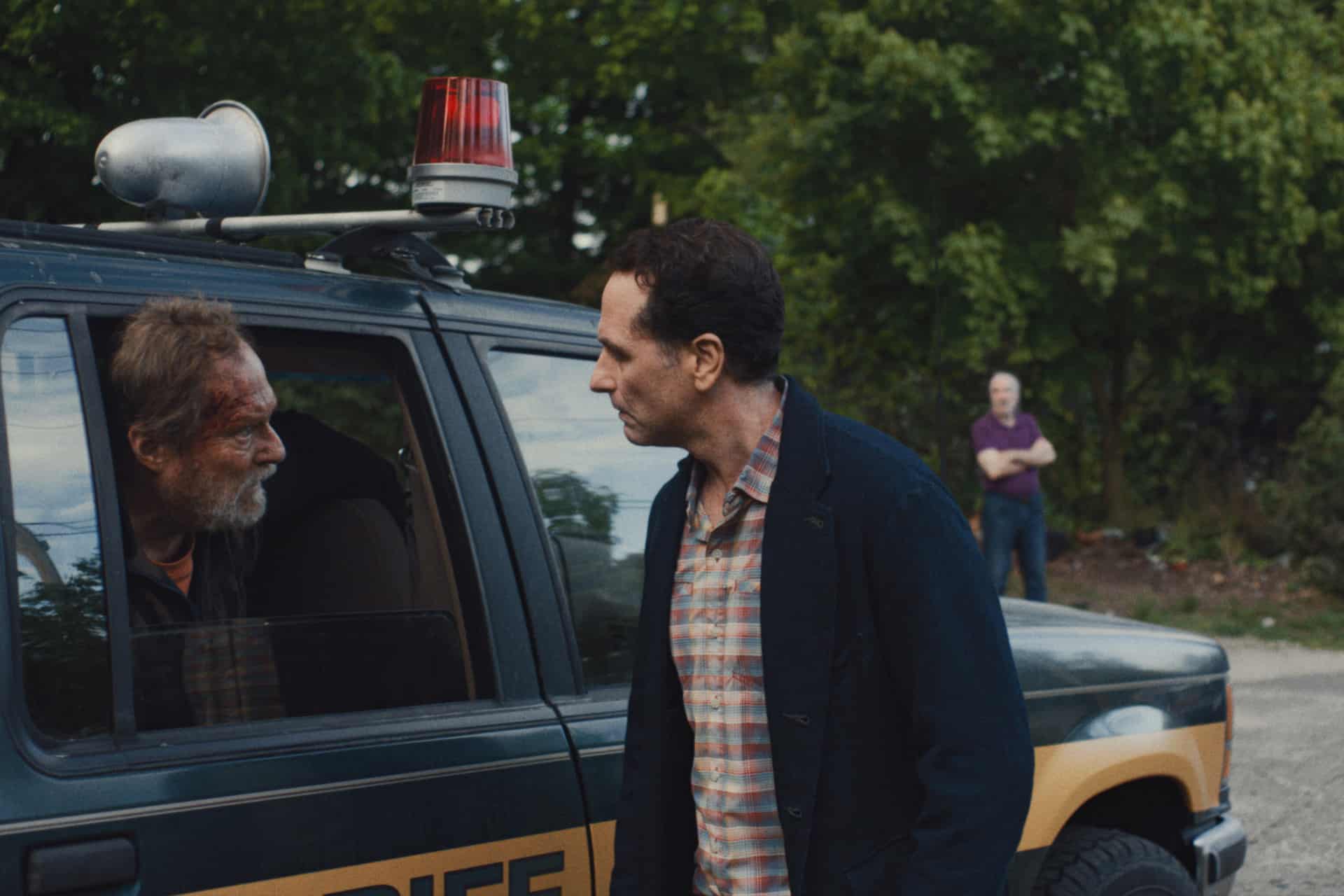 A wounded, disheveled man sits in the back of a sheriff’s vehicle while another man in a checked shirt stands outside talking to him. A third man stands in the background, surrounded by trees in this tense Apple TV Widow’s Bay scene.