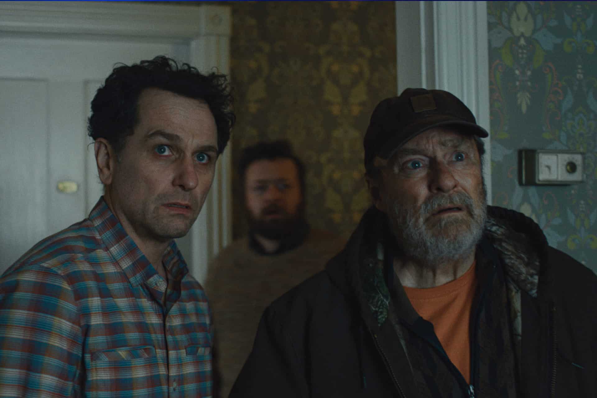 Three men stand indoors in patterned wallpaper, looking intently in the same direction, surprised or concerned. One wears a plaid shirt, another a dark jacket and cap, with a bearded man behind—they could be characters from Apple TV’s Widow’s Bay.