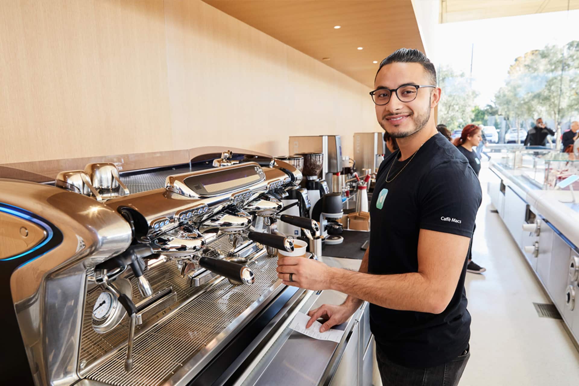 Apple Park Visitor Center - A barista wearing glasses and a black t-shirt smiles while preparing a coffee at a modern espresso machine inside the bright, contemporary Apple Park Visitor Center café.