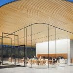 A modern building with large glass walls and a curved wooden ceiling at the Apple Park Visitor Center; people are sitting at tables inside while others stroll on a spacious, open plaza outside.