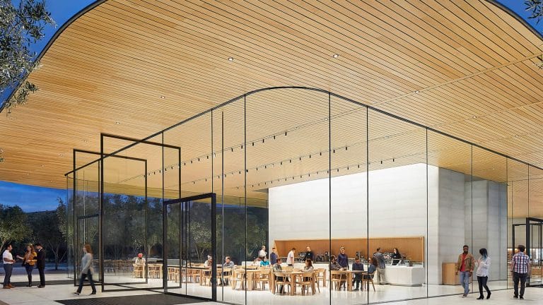 A modern building with large glass walls and a curved wooden ceiling at the Apple Park Visitor Center; people are sitting at tables inside while others stroll on a spacious, open plaza outside.