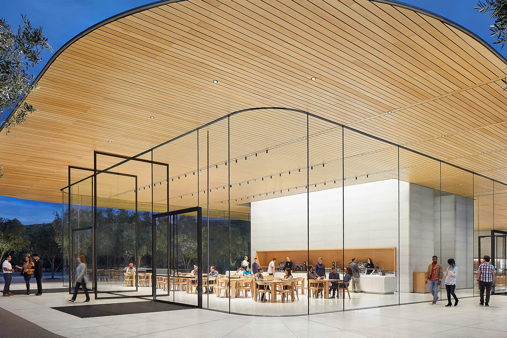 A modern building with large glass walls and a curved wooden ceiling at the Apple Park Visitor Center; people are sitting at tables inside while others stroll on a spacious, open plaza outside.