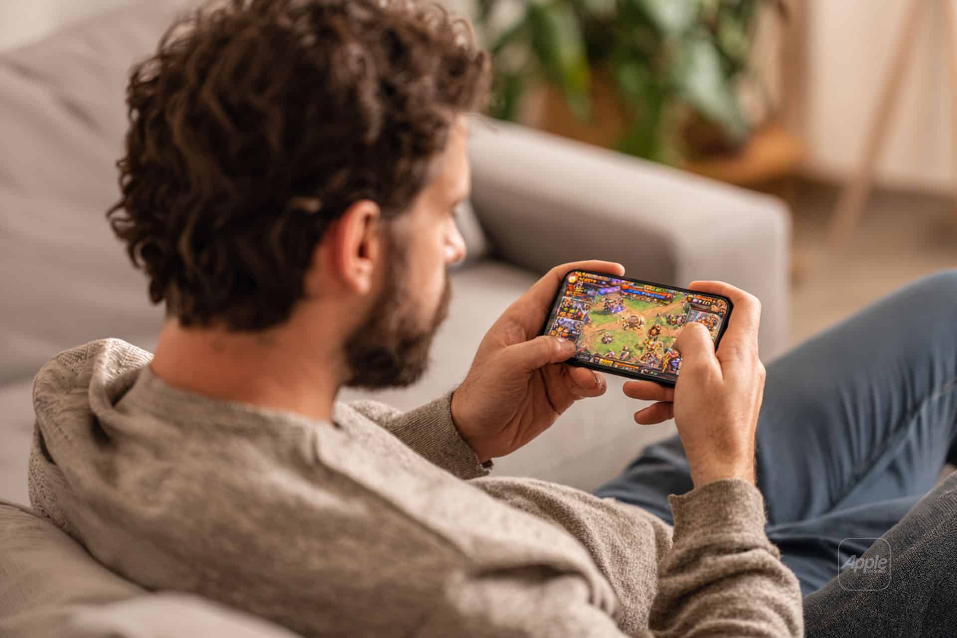 A man with curly hair sits on a couch, holding a smartphone in landscape mode and playing a colorful strategy game—a true digital hobbyist immersed in animated characters and exciting in-game elements.