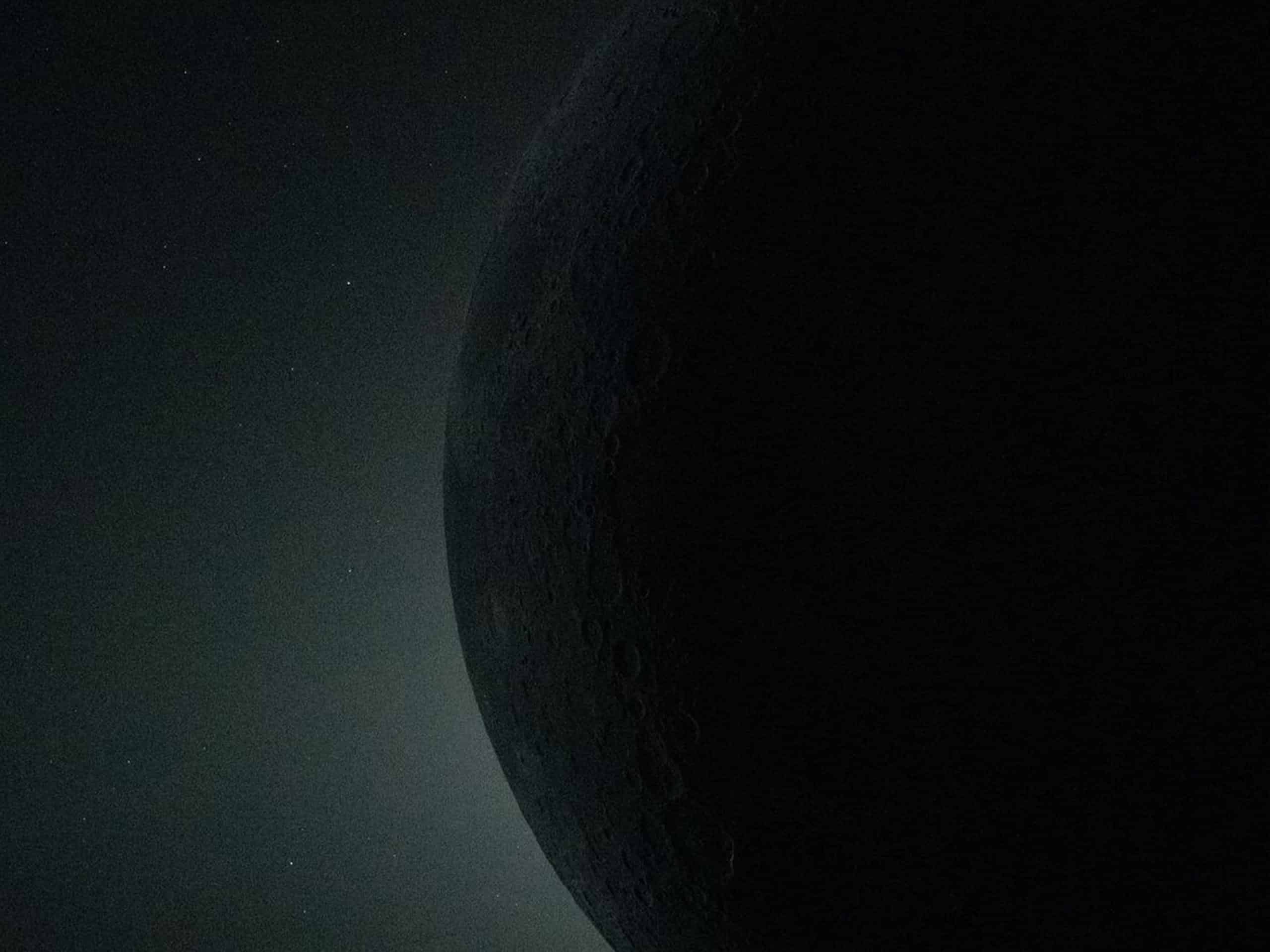 A shadowed, cratered surface of the Moon is seen from space, half illuminated and half in darkness, against a star-filled background—rendered with striking clarity as if powered by Apple GPU memory.