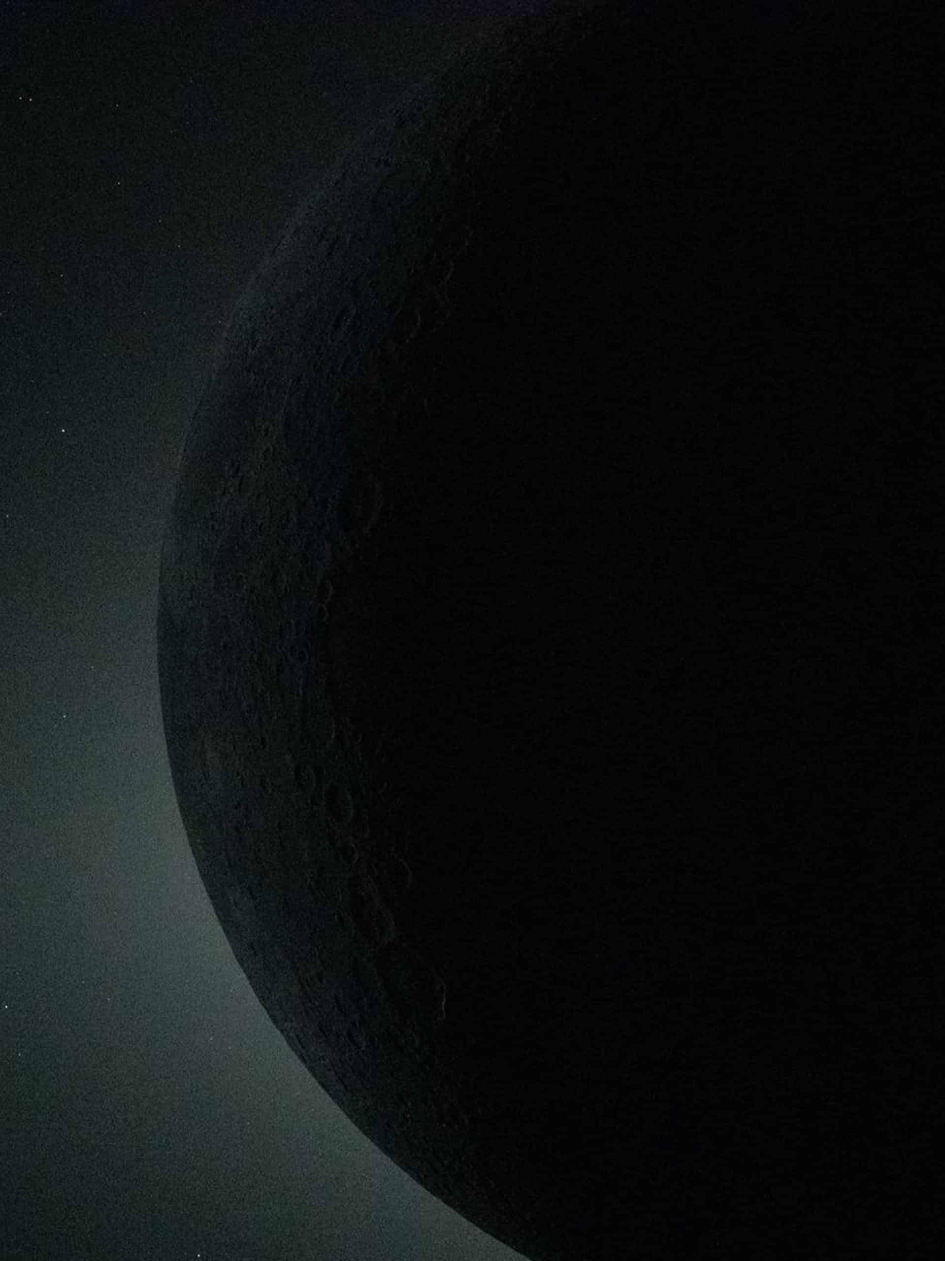 A shadowed view of the moon, with only a faint sliver of its surface illuminated, revealing subtle craters and textures against a dark, star-speckled background—rendered in crisp detail with Apple GPU memory.
