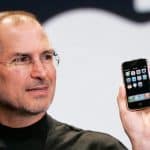 A man wearing glasses and a black turtleneck holds up an early model smartphone with app icons displayed on the screen, marking the start of the iPhone era, as he stands in front of a large, blurred logo in the background.
