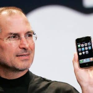 A man wearing glasses and a black turtleneck holds up an early model smartphone with app icons displayed on the screen, marking the start of the iPhone era, as he stands in front of a large, blurred logo in the background.