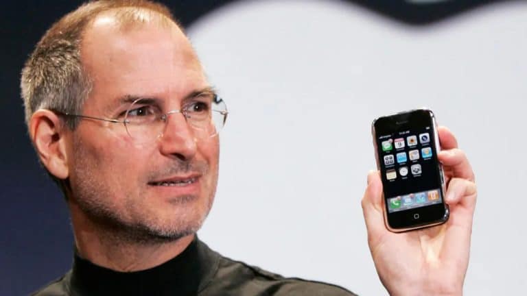 A man wearing glasses and a black turtleneck holds up an early model smartphone with app icons displayed on the screen, marking the start of the iPhone era, as he stands in front of a large, blurred logo in the background.