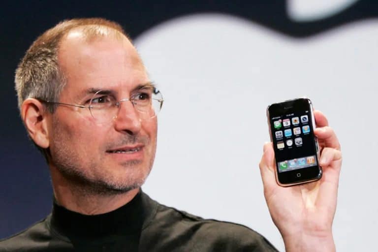 A man wearing glasses and a black turtleneck holds up an early model smartphone with app icons displayed on the screen, marking the start of the iPhone era, as he stands in front of a large, blurred logo in the background.