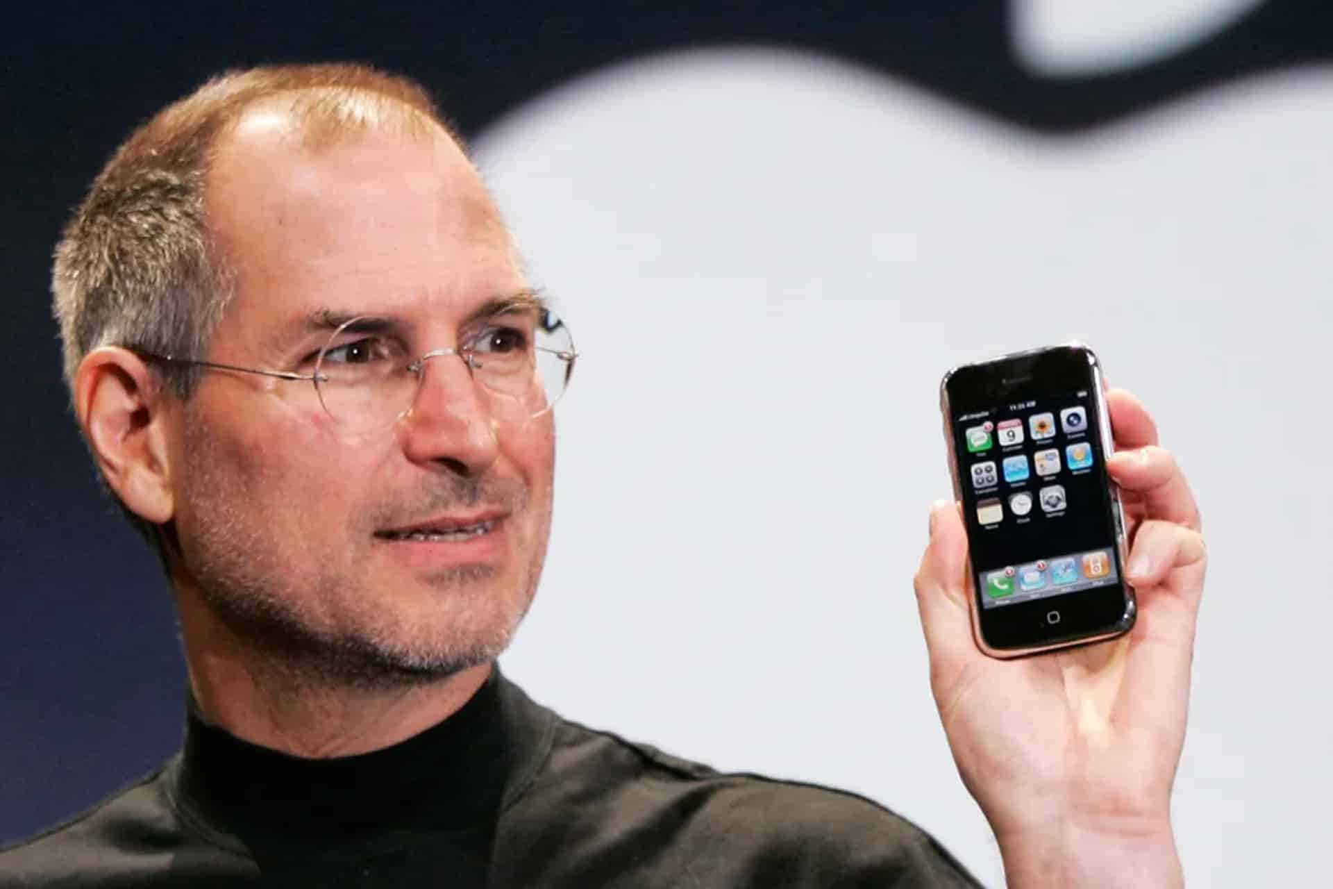 A man wearing glasses and a black turtleneck holds up an early model smartphone with app icons displayed on the screen, marking the start of the iPhone era, as he stands in front of a large, blurred logo in the background.