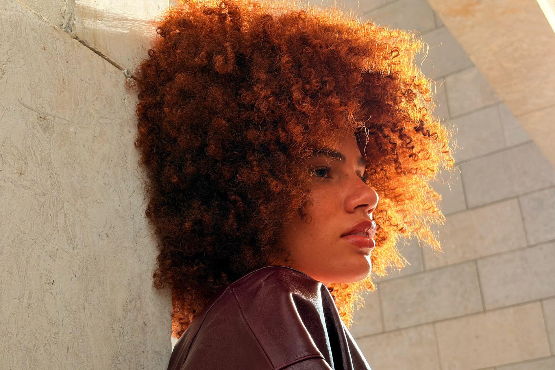 A person with voluminous, curly auburn hair stands against a light stone wall, sunlight from the iPhone Air camera illuminating their hair and face. They wear a dark top and are gazing thoughtfully into the distance.