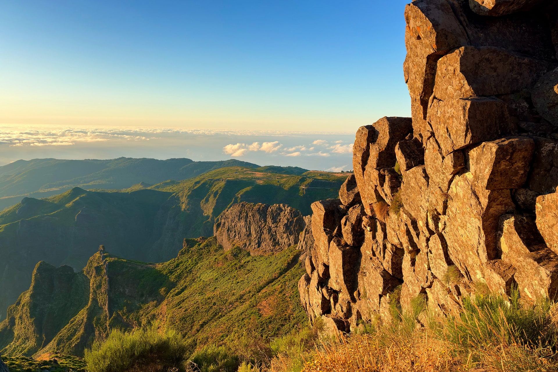 Rocky cliff in the foreground with sunlit, green mountains and valleys stretching into the distance, all beautifully captured using the iPhone Air camera under a clear blue sky and a layer of clouds on the horizon.
